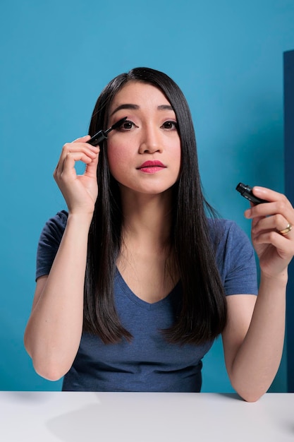 Porträt einer asiatischen Influencerin, die sich vor der Kamera mit Mascara schminkt, während sie ein Beauty-Tutorial für einen Vlogging-Kanal aufzeichnet. Künstlerischer Social-Media-Ersteller filmt Kosmetikrezension