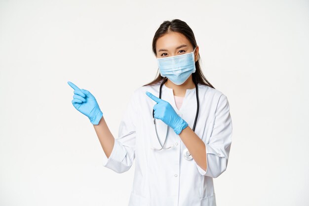 Porträt einer asiatischen Medizinerin, die mit den Fingern nach links zeigt, Gesichtsmaske und Gummihandschuhe trägt und in Klinikuniform auf weißem Hintergrund steht