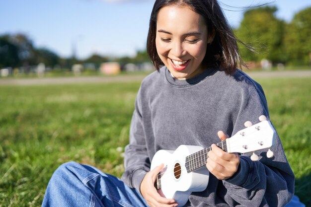 Porträt einer asiatischen Studentin, die Ukulele spielt und im Park singt, allein sitzend auf Decke und Enjo