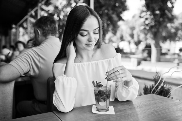 Porträt einer atemberaubenden jungen Frau, die mit Mojito-Cocktail im Café neben dem Park posiert Schwarz-Weiß-Foto