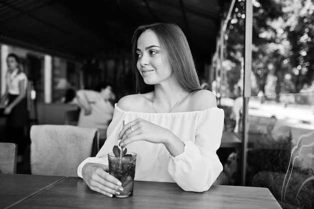 Porträt einer atemberaubenden jungen Frau, die mit Mojito-Cocktail im Café neben dem Park posiert Schwarz-Weiß-Foto