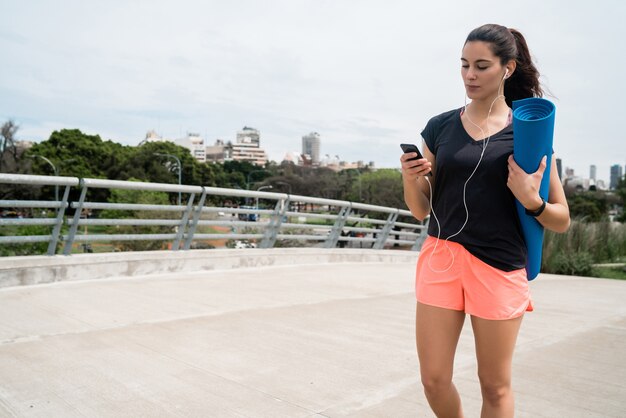 Porträt einer athletischen Frau, die auf der Straße geht, die eine Trainingsmatte hält, während Musik hört. Sport- und Lifestyle-Konzept.