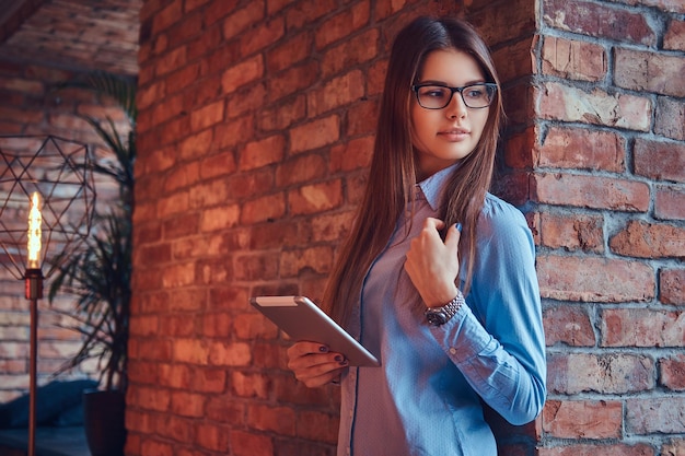 Porträt einer attraktiven, charmanten Geschäftsfrau mit Brille und blauem Hemd hält ein Tablet, das sich an einer Ziegelwand in einem Raum mit Loft-Design lehnt.