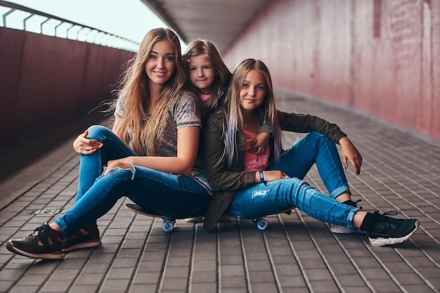 Porträt einer attraktiven Familie. Mutter und ihre Töchter sitzen zusammen auf einem Skateboard an einem Brückenfußweg.