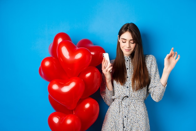 Porträt einer attraktiven jungen Frau, die Smartphone hält und in der Nähe von roten Ballons des Valentinsgrußes tanzt, stehend auf blauem Hintergrund