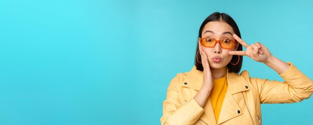 Porträt einer attraktiven Koreanerin mit Sonnenbrille, die ein Friedenszeichen in der Nähe von Augen zeigt, die Lippen küssen und Geste über blauem Hintergrund stehen