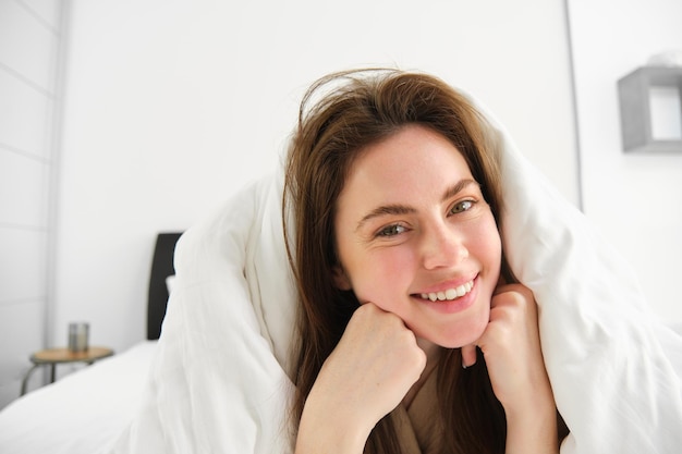 Porträt einer attraktiven lächelnden Frau, die entspannt im Bett liegt, bedeckt mit weißen Laken und einer warmen Bettdecke