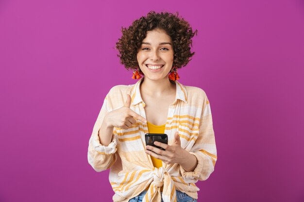 Porträt einer attraktiven lächelnden jungen frau mit lockigen brünetten haaren, die isoliert über einer violetten wand steht und auf das handy zeigt