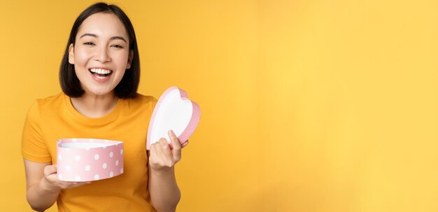 Porträt einer aufgeregten asiatischen Frau mit offenem Geschenkkarton und überraschtem, glücklichem Gesicht, das über gelbem Hintergrund steht