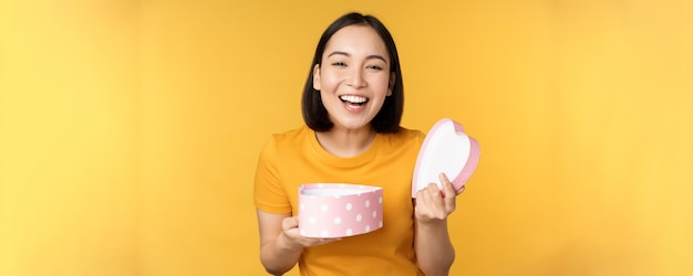 Porträt einer aufgeregten asiatischen Frau mit offenem Geschenkkarton und überraschtem, glücklichem Gesicht, das über gelbem Hintergrund steht