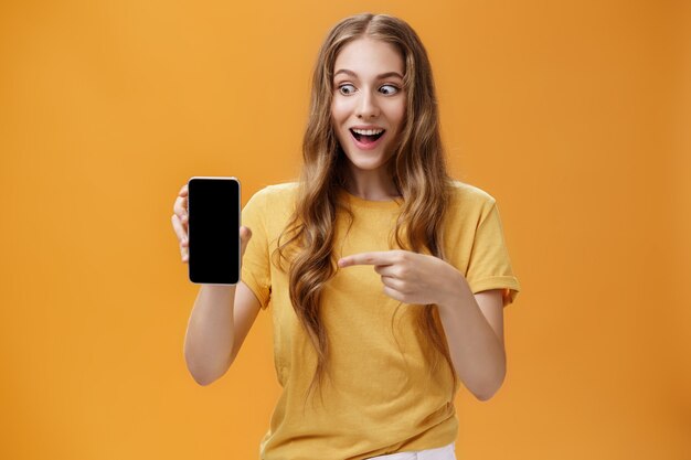 Porträt einer aufgeregten Frau, die sich erstaunt fühlt, ein tolles neues Smartphone in der Hand zu halten und auf den Handy-Bildschirm zu zeigen, der wie verrückt auf das Gerät knallt, das von einem coolen Technologieprodukt bezaubert wird.