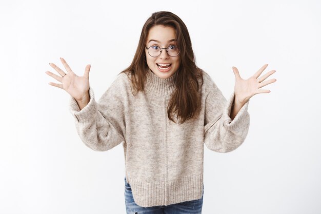 Porträt einer aufgeregten glücklichen jungen überraschten Frau in Brille und Pullover, die mit erhobenen Handflächen gestikuliert