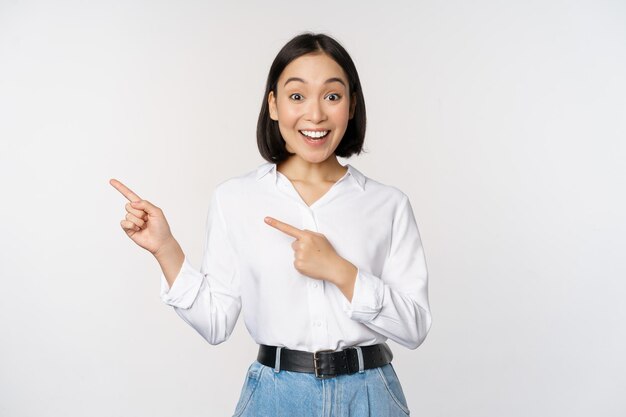 Porträt einer aufgeregten jungen asiatischen Bürodame, die mit dem Finger nach links auf den Rabatt zeigt, der die Verkaufsfahne zeigt, die über weißem Hintergrund steht