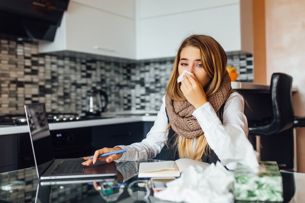 Porträt einer besorgten Geschäftsfrau, die Servietten hält und zu Hause in der Küche niest und einen Laptop benutzt