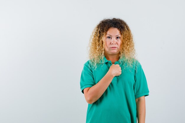 Porträt einer blonden Frau mit lockigem Haar, die im grünen T-Shirt die Hand über die Brust hält und verwirrte Vorderansicht schaut