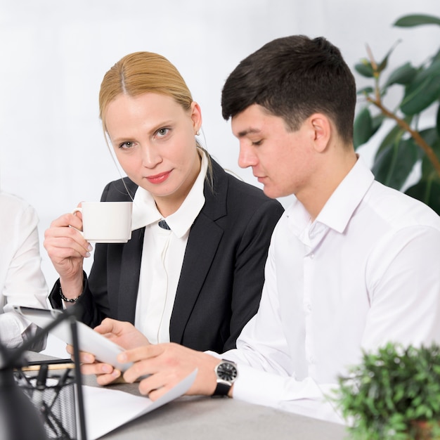 Porträt einer blonden jungen Geschäftsfrau, die in der Hand die Kaffeetasse sitzt mit dem männlichen Kollegen verwendet digitale Tablette hält