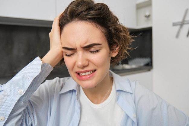 Porträt einer brünetten Frau, die ihren Kopf berührt, vor Schmerzen eine Grimasse zieht, Kopfschmerzen und schmerzhafte Migräne hat