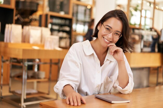 Porträt einer brünetten Frau im Café, die mit Notizbuch-Arbeitsunterlagen sitzt und auf Kamera und SMI blickt