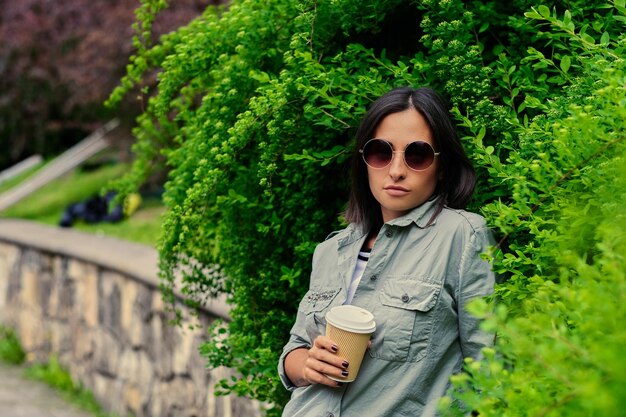 Porträt einer brünetten Frau mit Sonnenbrille trinkt Kaffee unterwegs in einem Sommerpark.