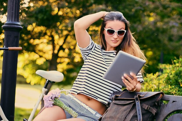Porträt einer brünetten Frau mit Tablet-PC mit Stadtfahrrad in einem Park im Hintergrund.