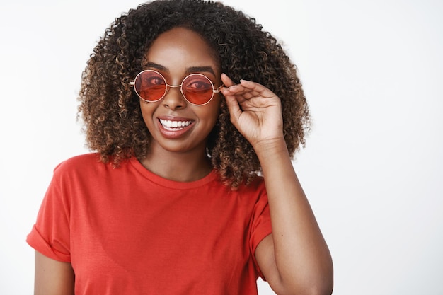 Porträt einer charmanten, unbeschwerten, enthusiastischen afroamerikanischen Frau mit lockigem Haarschnitt, die eine stilvolle rote Sonnenbrille trägt und optimistisch blickt