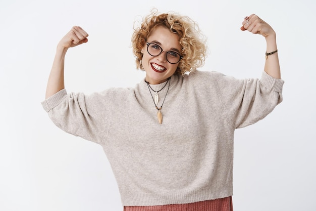 Kostenloses Foto porträt einer enthusiastischen glücklichen und selbstbewussten europäischen blonden frau in brille und pullover, die muskeln zeigt und freudig lächelt und kraft und mut über weiße wand zeigt