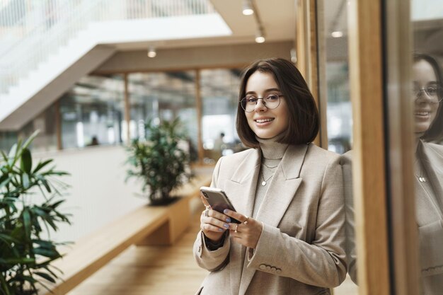 Porträt einer erfolgreichen, glücklichen jungen Frau, die eine prominente Karriere beginnt, die eine Nachricht sendet, ein Mobiltelefon verwendet und in die Kamera lächelt, während sie auf jemanden in der Rezeption der Bürohalle wartet, der Geschäftspartner trifft