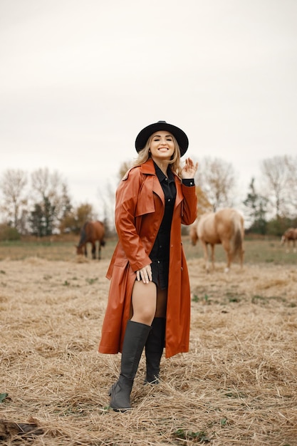 Porträt einer Frau auf einem Feld mit Pferden. Blonde Frau, die auf dem Feld steht, in die Kamera schaut und für ein Foto posiert. Frau mit schwarzem Kleid, rotem Ledermantel und Hut.