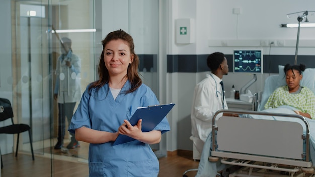 Porträt einer Frau, die als Krankenschwester arbeitet und Kontrollpapiere hält, während sie in der Krankenstation steht. Arzthelferin mit Dokumenten, die die Kamera betrachten und lächeln und dem Spezialisten helfen.