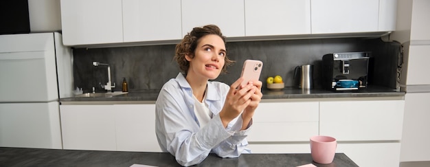 Porträt einer Frau, die denkt, während sie ein Smartphone in der Hand hält und entscheidet, was sie auf einer Handy-App bestellen soll