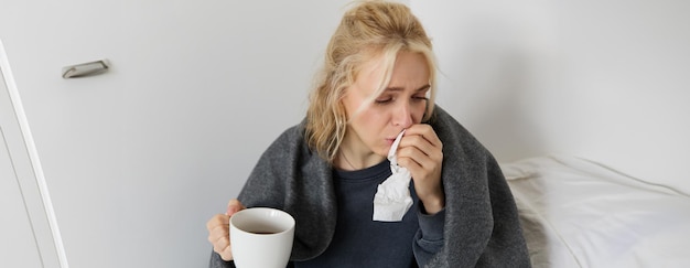 Porträt einer Frau, die sich erkältet, krank zu Hause bleibt, Tee trinkt, in eine Serviette niest, die Nase bläst