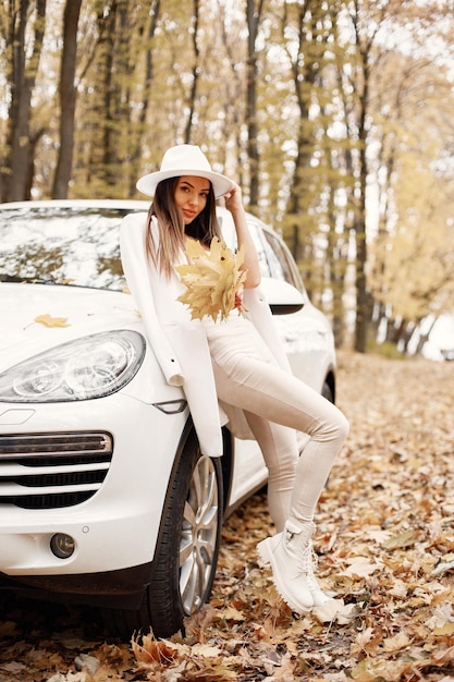 Porträt einer Frau im Herbstwald, die neben ihrem weißen Auto steht. Brunettefrau, die für ein Foto aufwirft. Frau mit weißem Hut, rosafarbenem T-Shirt und Hose.