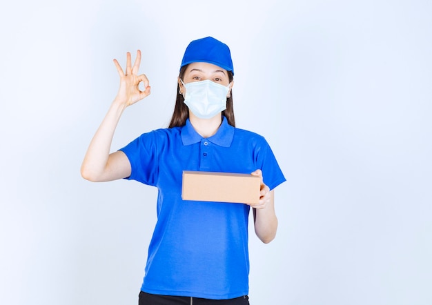 Porträt einer Frau in medizinischer Maske mit Pappschachtel, die eine ok Geste zeigt