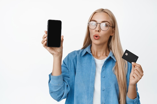 Porträt einer Frau mit Brille, die neugierig aussieht und eine interessante App auf dem Handy und Kreditkartenkonzept des Online-Shoppings und des kontaktlosen Bezahlens auf weißem Hintergrund zeigt