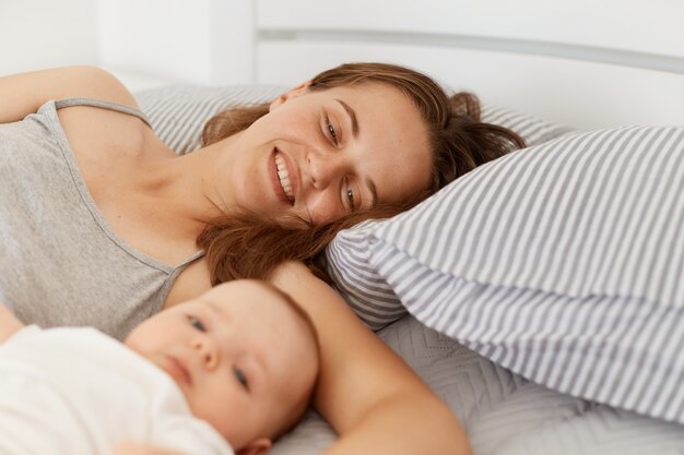 Porträt einer Frau mit Säuglingsbaby oder -junge, die am frühen Morgen im hellen Raum im Bett liegt, das Wochenende genießt und Zeit zusammen verbringt, glückliche Elternschaft.