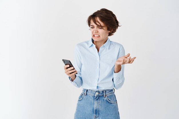 Porträt einer Frau zuckt zusammen und schaut angewidert auf den mobilen Bildschirm, der etwas Ekelhaftes mit Abneigung vor weißem Hintergrund liest
