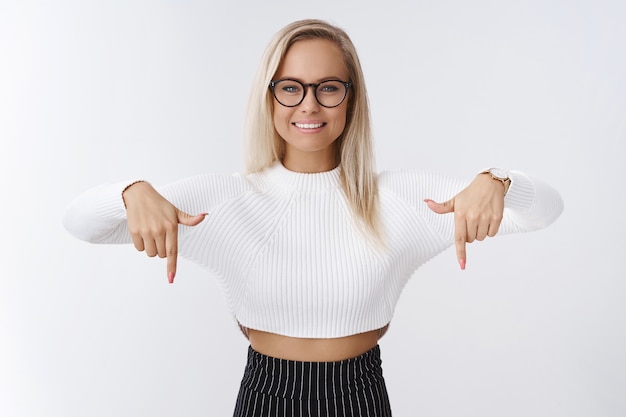 Porträt einer freundlich aussehenden, angenehmen und hilfsbereiten jungen persönlichen Assistentin in Brille und stylischem, kurz geschnittenem Pullover, der die Arme nach unten zeigt, als lächelnder, erfreuter Empfehlungsplatz.