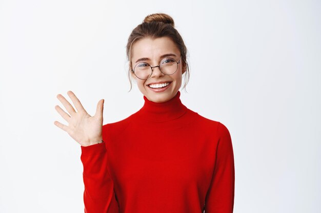 Porträt einer freundlichen CEO-Managerin, winkt mit der Hand und sagt Hallo, begrüßt Sie oder sagt auf Wiedersehen, macht Abschiedsgeste, steht in einer Brille auf Weiß
