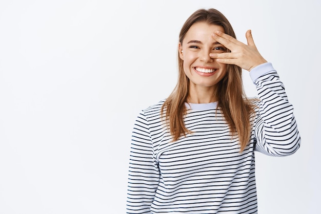 Kostenloses Foto porträt einer fröhlich lächelnden frau, die glück und freude ausdrückt, indem sie durch die finger über das auge schaut und von überglücklicher stellung vor weißem hintergrund grinst