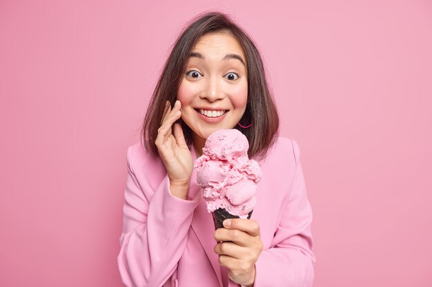 Porträt einer fröhlichen, brünetten jungen asiatischen frau, die große köstliche eiscreme hält, hat einen positiven ausdruck isst leckeren gefrorenen nachtisch nach der arbeit, gekleidet in formelle jacke, isoliert über rosa wand.