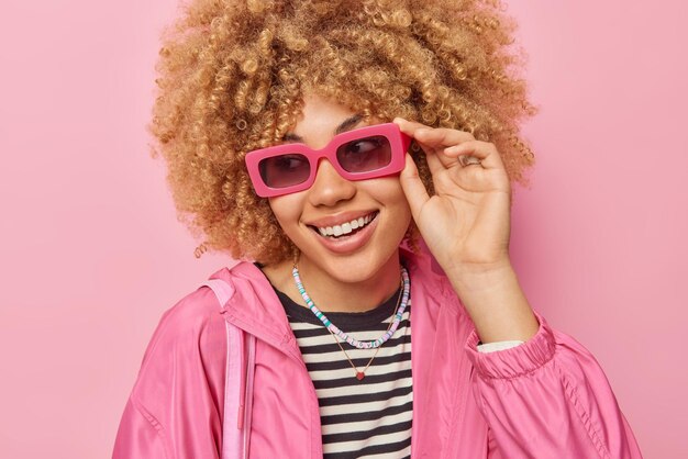 Porträt einer fröhlichen Frau mit lockigem, buschigem Haar, das sich konzentriert hat, trägt eine Sonnenbrille und eine Freizeitjacke, die gute Laune hat, isoliert auf rosafarbenem Hintergrund Emotionen und Stil