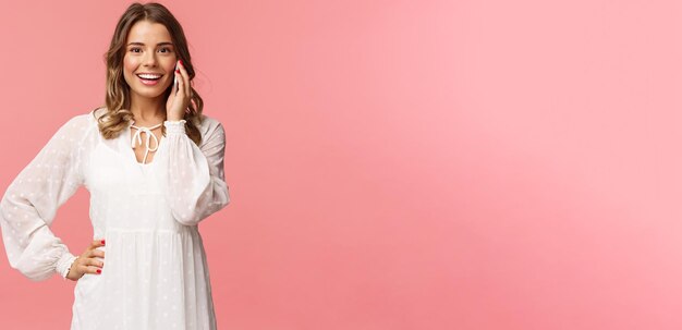 Porträt einer fröhlichen, gutaussehenden blonden frau in weißem kleid, die am telefon spricht, hält smartphone in der nähe des ohrs und schaut optimistisch lächelnd in die kamera, bespricht freunde mit date-stand auf rosa hintergrund