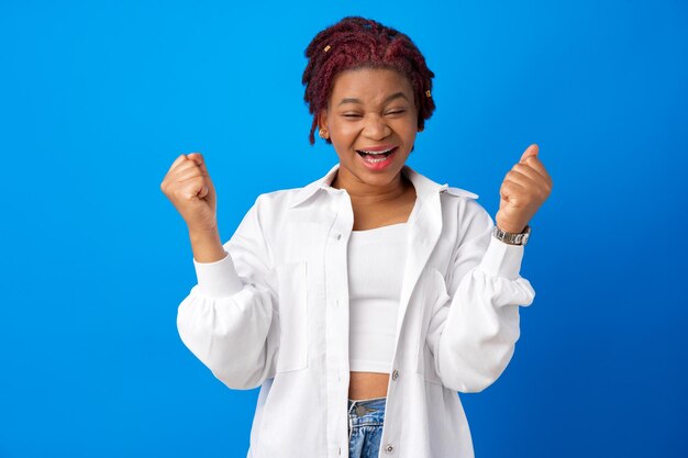 Porträt einer fröhlichen jungen Afrikanerin mit erhobenen Händen vor blauem Hintergrund