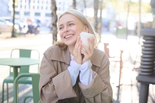 Kostenloses Foto porträt einer fröhlichen jungen blonden frau, die es genießt, in einem café im freien kaffee zu trinken und