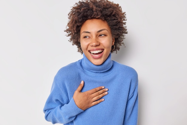 Porträt einer fröhlichen, lockigen Frau, die breit lächelt, hält die Hand auf der Brust und kann nicht aufhören, über lustigen Witz zu lachen, trägt einen lässigen blauen Pullover einzeln auf weißem Hintergrund. Positives Konzept der menschlichen Emotionen