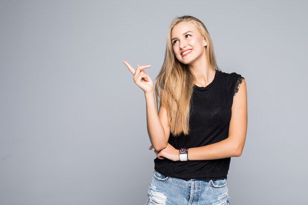 Porträt einer fröhlichen und glücklichen jungen schönen Frau in einem schwarzen T-Shirt, das mit einem Lächeln in die Kamera schaut und einen Finger auf die Seite zeigt, die auf dem grauen Hintergrund lokalisiert wird.