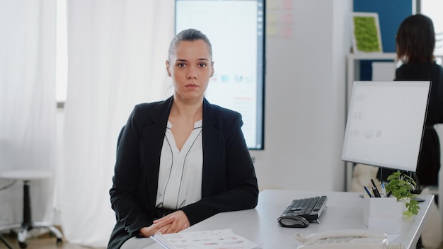 Porträt einer Geschäftsfrau mit Bürojob, die am Schreibtisch mit Computermonitor und Datendiagrammen auf dem Bildschirm sitzt. Unternehmensangestellter blickt in die Kamera und bereitet sich auf die Arbeit an Geschäftsprojekten vor