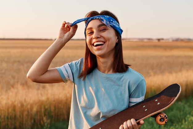 Porträt einer glücklich lachenden Frau, die im Sommer im Freien posiert, Skateboard in den Händen hält, ihr Haarband berührt und die Kamera mit positiven Emotionen betrachtet.