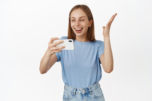 Porträt einer glücklich lächelnden Frau, die sich etwas auf dem Handy ansieht, sich freut, im Smartphone-Videospiel gewinnt, fröhlich auf den Bildschirm schaut und gegen weiße Wand steht