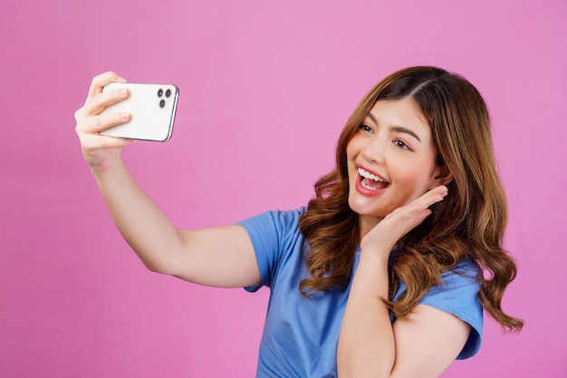 Porträt einer glücklich lächelnden jungen Frau, die ein lässiges T-Shirt-Selfie mit Smartphone trägt, das über rosafarbenem Hintergrund isoliert ist